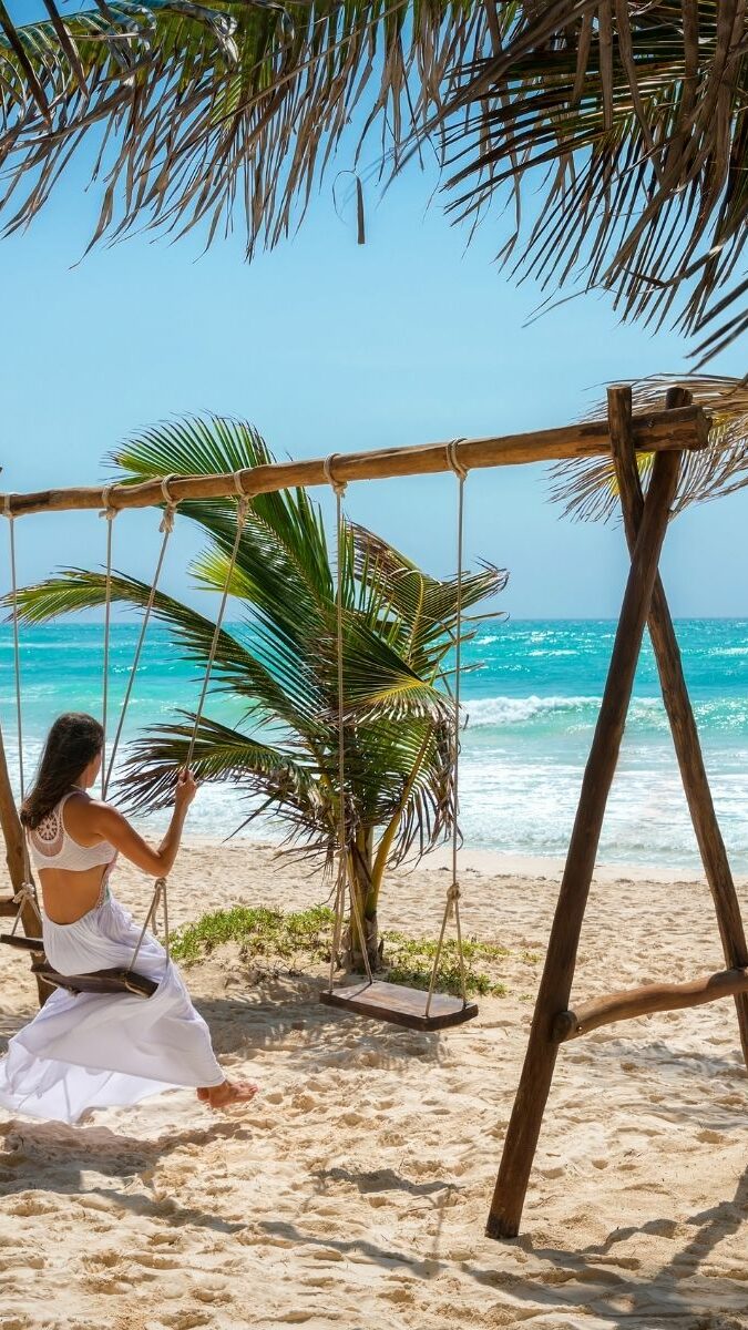 Femme assise sur une balançoire en bois sur une plage de sable blanc bordée de palmiers à Tulum