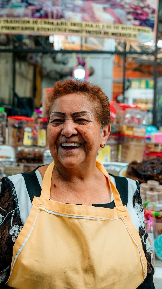 Marchande souriante dans son tablier jaune au marché d'Oaxaca entourée de produits locaux